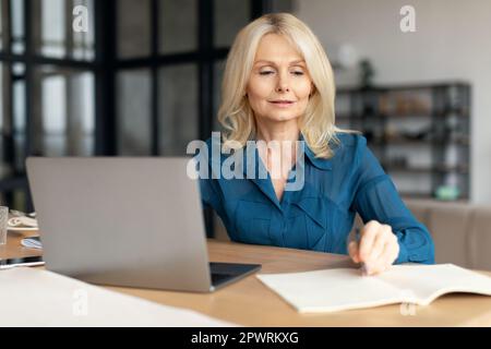 Mature blonde lady working on laptop at home office, senior woman taking notes during online work meeting or webinar Stock Photo