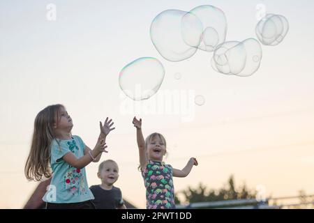The happy children are playing with soap bubbles outdoors on sky background. Stock Photo