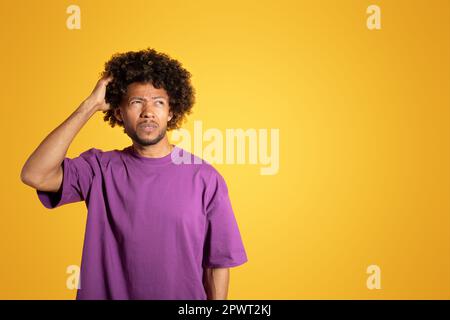 Pensive adult black curly man in purple t-shirt scratching his head, think, look at free space Stock Photo