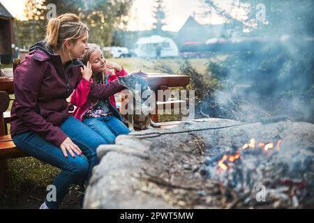 Family with dog sitting at campfire during summer vacation. Mother and daughter spending time on summer weekend. Vacation trip close to nature Stock Photo