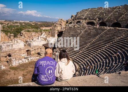 A tourist couple sit at the top of the well-preserved amphitheatre in the ancient Roman city of Side in Antalya province, Turkey (Turkiye). The Roman Stock Photo