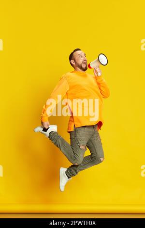 Full-length portrait of bearded man, influencer, jumping and shouting in megaphone against vivid yellow studio background Stock Photo