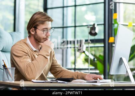 A young businessman sits in the office at a table, works with documents, looks at notes in a notebook, thinks about a strategy, analyzes. He rubs his beard thoughtfully with his hand. Stock Photo