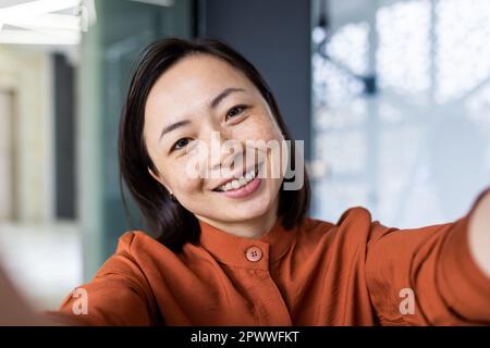 Young beautiful Asian business woman taking selfie photo, female worker talking on video call looking into smartphone camera smiling and happy, inside office building at workplace. Stock Photo