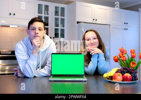 teenagers near laptop with green screen chromakey in kitchen advertising food goodies cooking show training online holiday day off boy and girl 13-16 years old leaned on table look frame real people Stock Photo