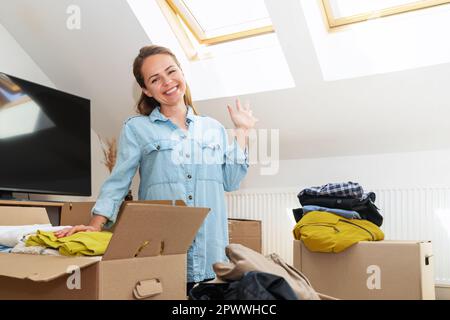 Woman unpacking her clothing smiling and waving hand. Moving house concept. Stock Photo