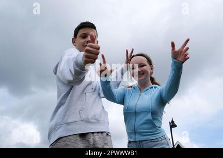couple of teenagers a boy and a girl stand against the background of a sky with clouds raise their thumbs up build horns to each other spread their fingers laughing having fun having fun approving Stock Photo