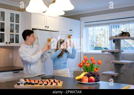 Happy interracial couple dancing in the kitchen, singing while cooking breakfast or dinner, young woman and a handsome man smiling and having fun together, holding kitchen utensils Stock Photo