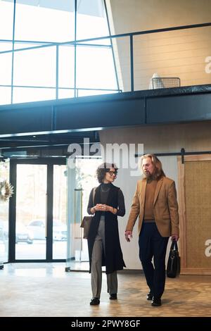 Two mature colleagues in quiet luxury attire discussing working points while walking along spacious corridor or lounge in business center Stock Photo