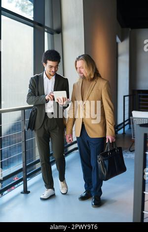 Two intercultural businessmen in formalwear discussing online financial data in tablet while walking along spacious corridor of office center Stock Photo