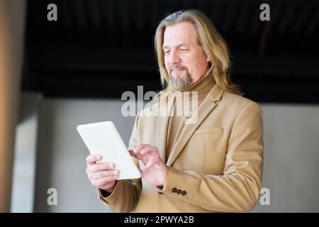 Elegant mature businessman in beige formalwear using tablet while analyzing online graphic data or entering points of new project Stock Photo