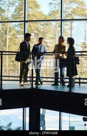 Working meeting of four intercultural employees in formalwear standing against large window in corridor of office center and brainstorming Stock Photo