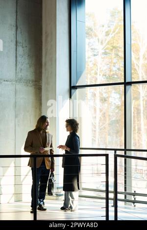 Outlines of two mature colleagues discussing project points while standing in sunlit corridor of office center between window and railing Stock Photo