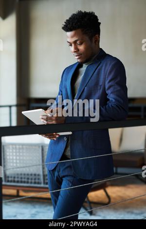 Young serious African American businessman in dark blue suit using tablet while standing in corridor of office center and analyzing online data Stock Photo