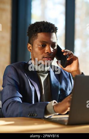 Young African American male broker talking to client on smartphone and using laptop while analyzing exchange rates on stock market Stock Photo