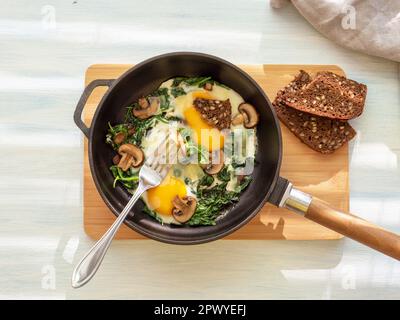 Idea for breakfast. Fried eggs with spinach, mushrooms, cheese and bread in cooking pan. Healthy homemade dish for low carb diet on a light blue Stock Photo