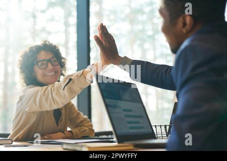 Focus on hands of two young intercultural successful colleagues giving each other high-five while sitting by workplace in boardroom Stock Photo