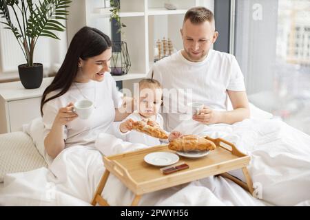 Happy husband and wife holding coffee cups while cute kid reaching out for delicious food on bed tray in modern studio flat. Small caucasian family having enjoyable meal together in home interior. Stock Photo