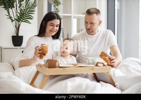 Cheerful caucasian parents in casual clothes sharing mealtime with cute infant daughter while staying at home for morning on Sunday. Smiling adults and small kid having fun together at weekend. Stock Photo