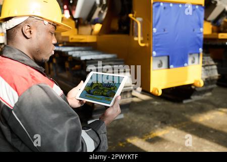 Side view of young African American engineer looking at machine picture on tablet screen while standing in workshop with industrial equipment Stock Photo