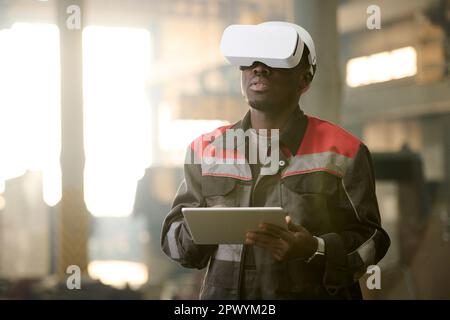 Young machine operator in vr headset and workwear using tablet while standing in front of machine and taking control of its work Stock Photo