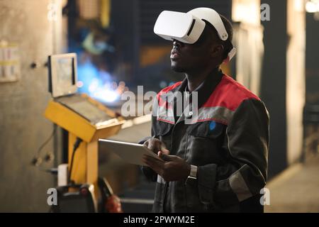 Young African American worker of factory in vr headset using tablet while operating industrial machine during technical production process Stock Photo