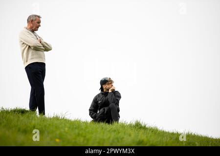 Two men look down from the top of a hill. One older man standing and a younger one on one knee. Stock Photo