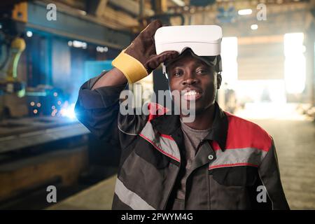 Happy young African American welder in workwear and protective gloves taking off vr headset and looking at camera against working machine Stock Photo