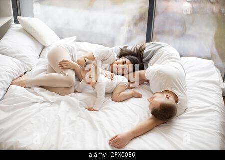 Top view of relaxed mature parents in leisurewear lying on soft bed while cute infant girl crawling on white blanket in home interior. Beautiful family of three enjoying free time indoors at weekend. Stock Photo