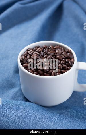 A view of a white mug full of dark roasted coffee beans on a wrinkled blue cloth surface. Stock Photo