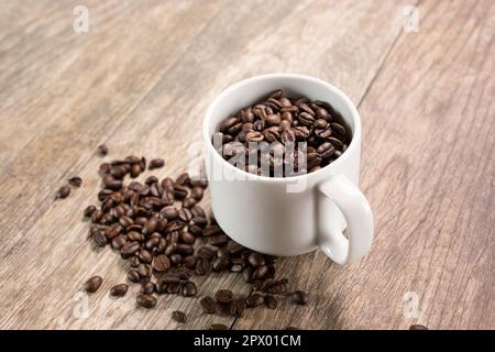 A view of a white mug filled with coffee beans, with a pile of coffee beans below in a mess on the table. Stock Photo