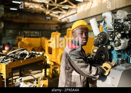 Young African American engineer in workwear and yellow protective helmet looking at camera while operating huge industrial machine Stock Photo