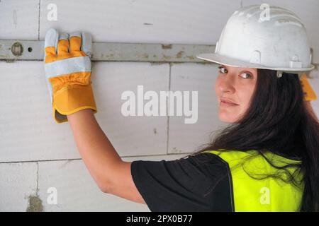 Young brunette woman construction worker in white hard hat and green high visibility vest, measuring wall with spirit bubble level, looking back over Stock Photo