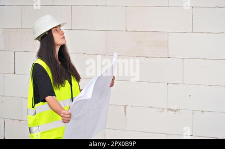 Young woman in white hard hat helmet and yellow green high visibility vest, standing next to construction wall, holding paper blueprints plans, lookin Stock Photo