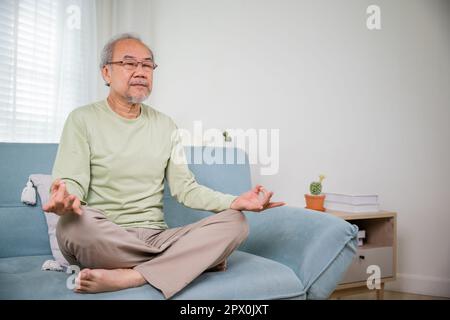 Lifestyle calm senior man lotus pose doing yoga for mental balance breathing air relaxing on sofa at home, Asian elderly old man sitting practise yoga Stock Photo