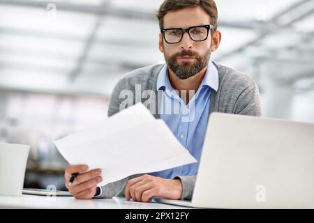 Getting the paperwork done. A young businessman going over paperowrk while working on his laptop in the office. Stock Photo