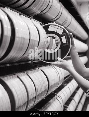 1980s HANDS OF COMPUTER TECHNICIAN SELECTING MEMORY STORAGE TAPE FROM LARGE RACK OF SIMILAR STORAGE TAPES - o4126 HAR001 HARS ROWS SIMILAR HIGH TECH WAREHOUSE ELECTRONIC REELS SOLUTIONS STORE ROOM VIDEO BLACK AND WHITE CAUCASIAN ETHNICITY COMPUTING HANDS ONLY HAR001 MAGNETIC OLD FASHIONED SELECTING TAPES Stock Photo