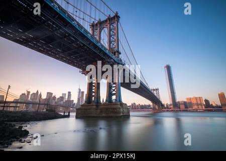 Manhattan Bridge seen from the coast next to the D.U.M.B.O area, during dusk with completely clear skies. Manhattan skyline can be seen on the back Stock Photo