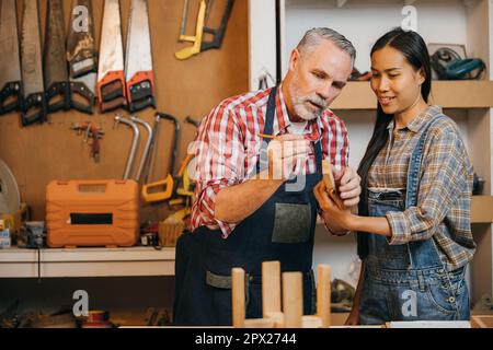 Craftsmen senior man teaching woman apprentice at woodshop, Carpenter training wood for female standing at wood table in workshop, National Carpenters Stock Photo