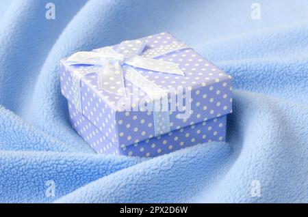 A small gift box in blue with a small bow lies on a blanket of soft and furry light blue fleece fabric with a lot of relief folds. Packing for a gift Stock Photo