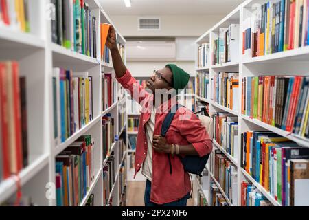 Nerd student african american man choosing book in university library taking it from shelf. Stock Photo