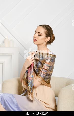 Sweet young woman with her shoulders wrapped in silk scarf sitting in armchair Stock Photo