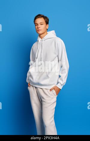 Young man wearing white sweatshirt and sweatpants standing over blue studio background with hands in pockets Stock Photo