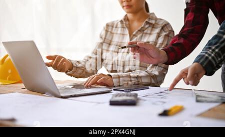 Cropped image of a female architect working with engineers in the office, checking plan on laptop and blueprint together. Stock Photo