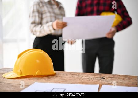 Close-up image of a yellow safety helmet hat and blueprint are on a table over blurred background of engineers. Stock Photo
