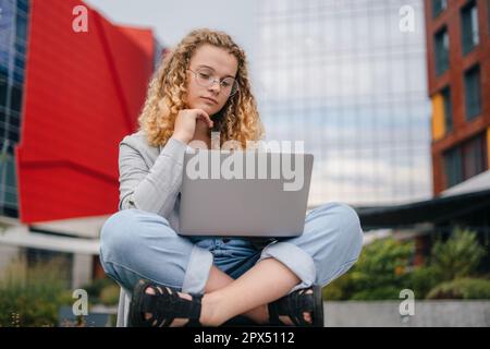 Portrait of beautiful stylish woman student wearing fashion clothes and eyeglasses, working remotely on a laptop on vacation. Woman surfing internet. Stock Photo