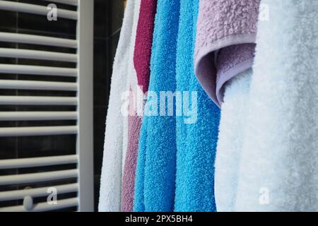 Towels hang next to a heated towel rail, wall radiator or radiator. White, blue, pink, red towels. Organization of household items in the bathroom. Ho Stock Photo