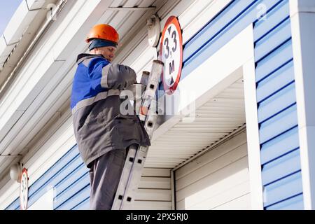 Male electrician stands on ladder and replaces light bulb in ceiling lamp on gate of industrial building or garage. Genuine workflow Stock Photo