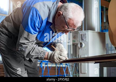 Elderly worker in glasses works in shop. Man in work clothes is bent over workbench in workshop and is making markings or drawings on metal sheet. Industrial background. Stock Photo