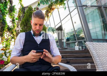 Young hipster in a white shirt, black vest looks into a smartphone while sitting in a penthouse Stock Photo
